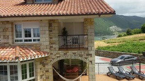 A two-story house with a tiled roof, stone walls, a balcony with a metal railing, and a view of a mountainous landscape. - Exclusive apartment in beach Sonabia with a view to the sea and the mountain. (Castro Urdiales)