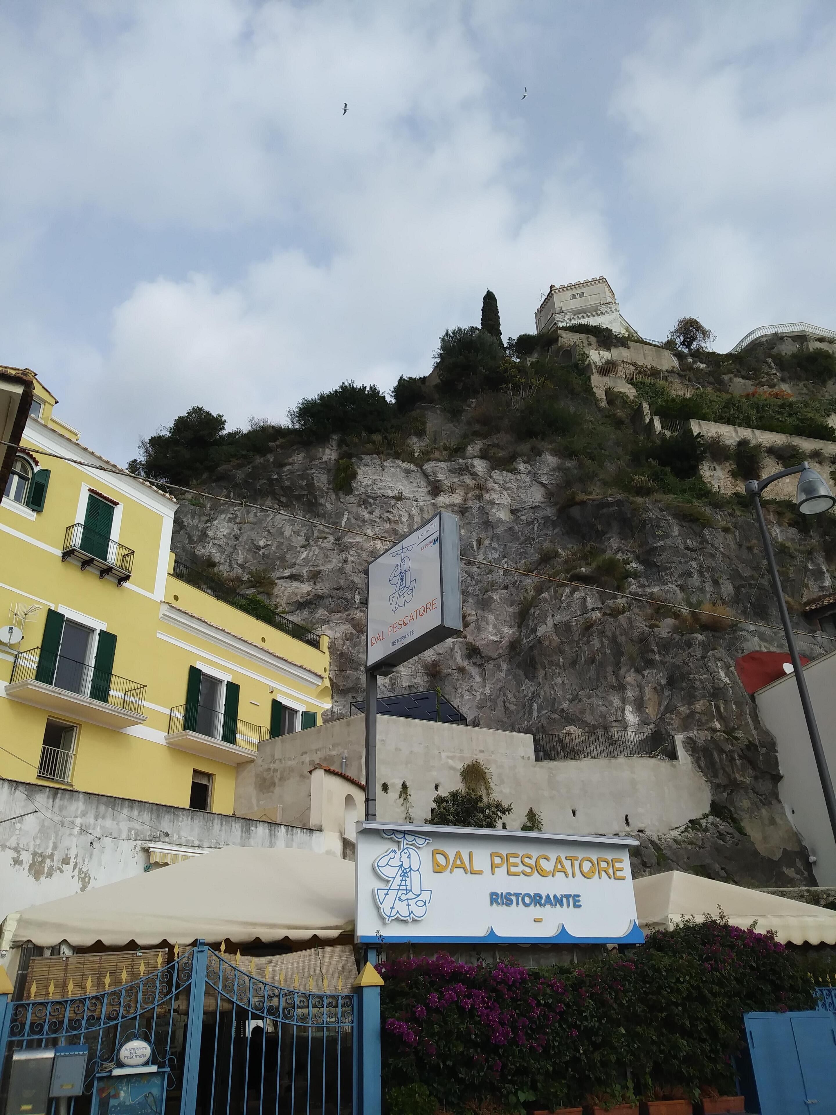 Um restaurante chamado DAL PESCATORE com uma placa, situado numa área costeira com edifícios e um penhasco rochoso ao fundo.