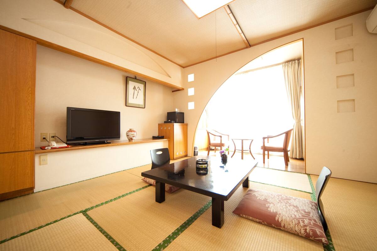 A traditional Japanese room with tatami flooring, a television on a low shelf, a wooden cabinet, and a sliding door leading to a balcony.
