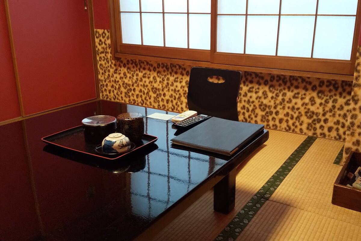 A traditional Japanese room with tatami flooring, a low table with a tea set, a black chair, and a window with a view of the outside.