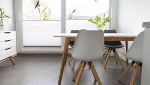 Un área de comedor moderna con una mesa y sillas blancas, ventanas grandes con persianas y vistas al exterior.