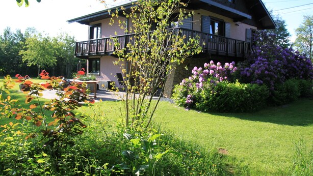 Une maison à deux étages dotée d’un balcon, entourée d’un jardin bien entretenu où poussent diverses plantes et fleurs.
