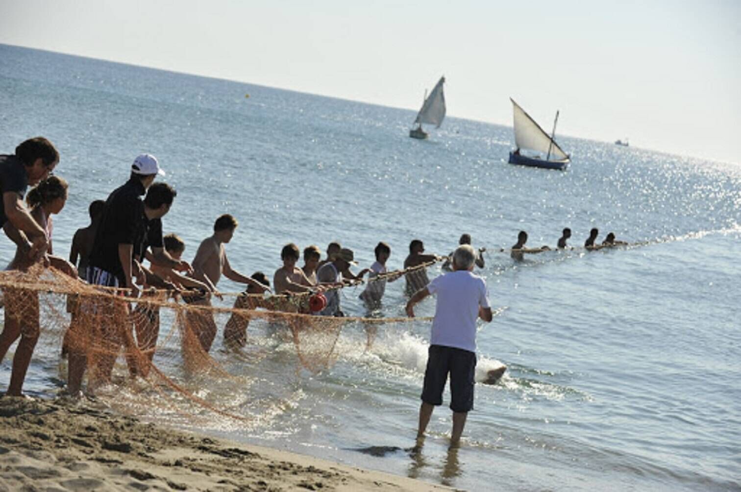 Escena de playa con gente pescando, barcos al fondo y cielo despejado.
