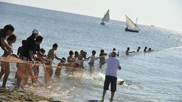 Escena de playa con gente pescando, barcos al fondo y cielo despejado.