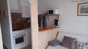 A compact kitchen with a microwave, coffee maker, and a hanging rack. A stone wall separates the kitchen from a living area with a sofa and pillows. - African reserve Sigean 15 mn, beach 200 m, Le Mieukerien (Port-La-Nouvelle)