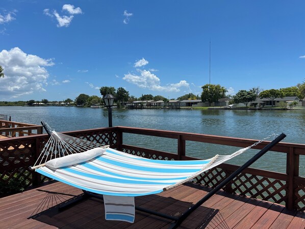 A hammock on a wooden deck overlooking a lake with houses in the background.