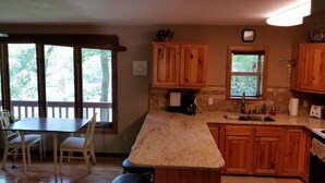 A kitchen with wooden cabinets, a granite countertop, a dining area with a table and chairs, and a view of trees through large windows. - Lakefront Adventure: Private Hot Tub,  Dock, Nearby Rafting, Biking & More! (Robbinsville)