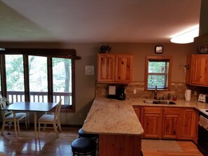 A kitchen with wooden cabinets, a granite countertop, a dining area with a table and chairs, and a view of trees through large windows. - Lakefront Adventure: Private Hot Tub,  Dock, Nearby Rafting, Biking & More! (Robbinsville)