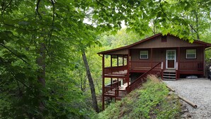 A cabin with a wooden deck and stairs leading to a front door, surrounded by dense green foliage. - Lakefront Adventure: Private Hot Tub,  Dock, Nearby Rafting, Biking & More! (Robbinsville)