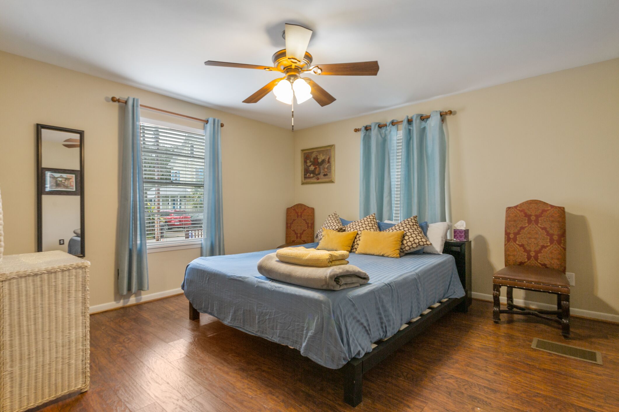 Une chambre à coucher comprenant un lit, une chaise, une table de chevet, un ventilateur de plafond et une fenêtre avec des rideaux.