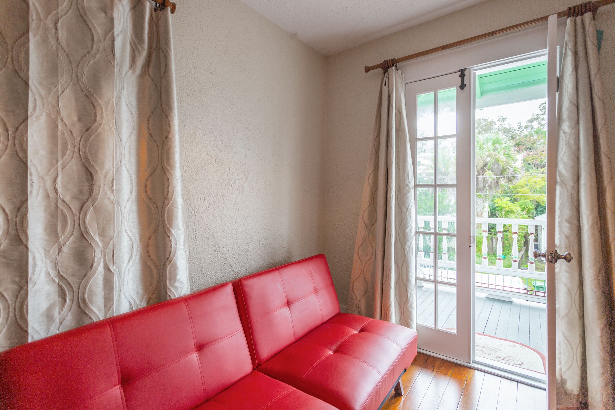 Um sofá vermelho numa sala com parede branca e porta de vidro deslizante que dá para área externa com deck de madeira e cerca branca.