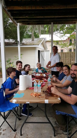 Un groupe de personnes s’est réuni autour d’une table où étaient disposés de la nourriture et des boissons. Elles étaient assises sur des chaises pliantes, à l’abri sous une zone couverte, avec une maison en arrière-plan.