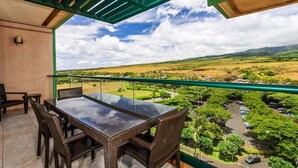 Un balcon avec une table en verre et des chaises en osier, donnant sur un paysage verdoyant avec une route et des voitures garées.