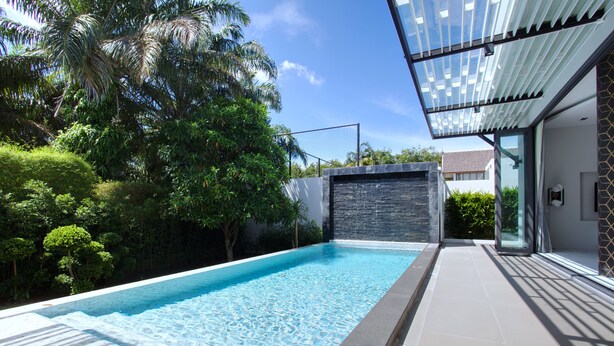 Ein moderner Pool mit Steinwand und Glasüren, umgeben von üppiger Vegetation und blauem Himmel.
