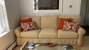 Un sofá color crema con almohadones estampados, una mesa de centro de vidrio con revistas, y una obra de arte enmarcada encima.