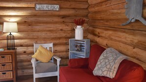 A cozy cabin interior with a wooden wall, a sign reading "Rustic Alaska", a white chair with a yellow pillow, a red sofa, and a small white vase with red flowers. - Comfy Cabin Nestled in the Woods (Houston)