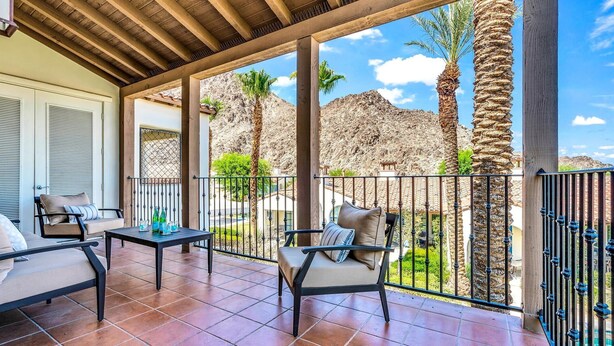 Un espacio de estar techado al aire libre con sillas y una mesa, con vistas a un paisaje rocoso con palmeras.