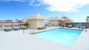 Une piscine sur le toit, dotée de chaises longues et d’un jacuzzi, le tout entouré d’une clôture et avec des bâtiments en arrière-plan.