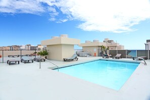 Piscina en la azotea con sillones de descanso y bañera de hidromasaje, rodeada por una valla y edificios.