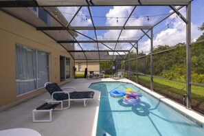 Piscina externa com espreguiçadeiras e boias, cercada por uma área coberta com vista para uma casa e vegetação.