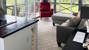 A spacious room with large windows, a ceiling fan, a red armchair, a grey sofa, and a dining area with a table and chairs.