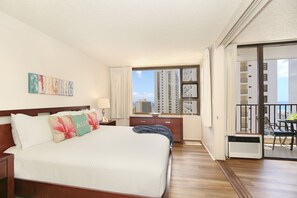A hotel room with a large bed, a nightstand, a dresser, a window with a view of tall buildings, and a balcony with a table and chairs. - Partial Ocean View, one-bedroom with AC, Wi-Fi, parking, short walk to beach! (Honolulu)