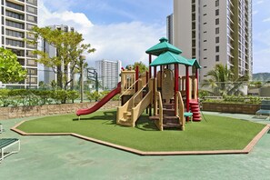A playground with slides and climbing structures, surrounded by a green artificial turf area, with tall residential buildings in the background.