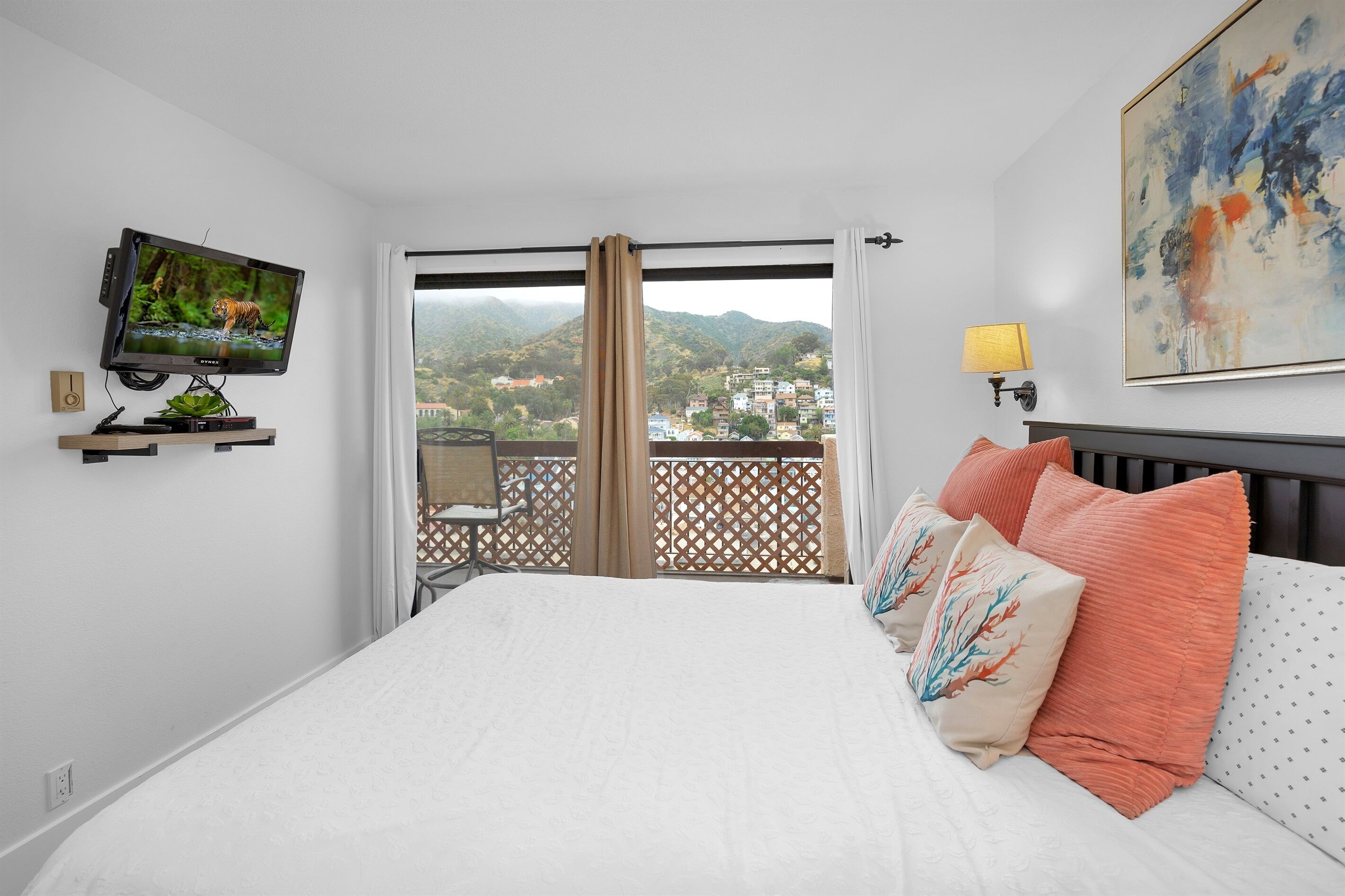 Een hotelkamer met een bed, een televisie op een standaard, een schilderij, een lamp en uitzicht op de bergen door het raam.