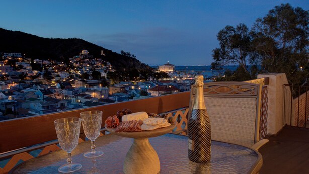 A table with a bottle of champagne, glasses, and a plate of food on a balcony overlooking a coastal town at dusk.