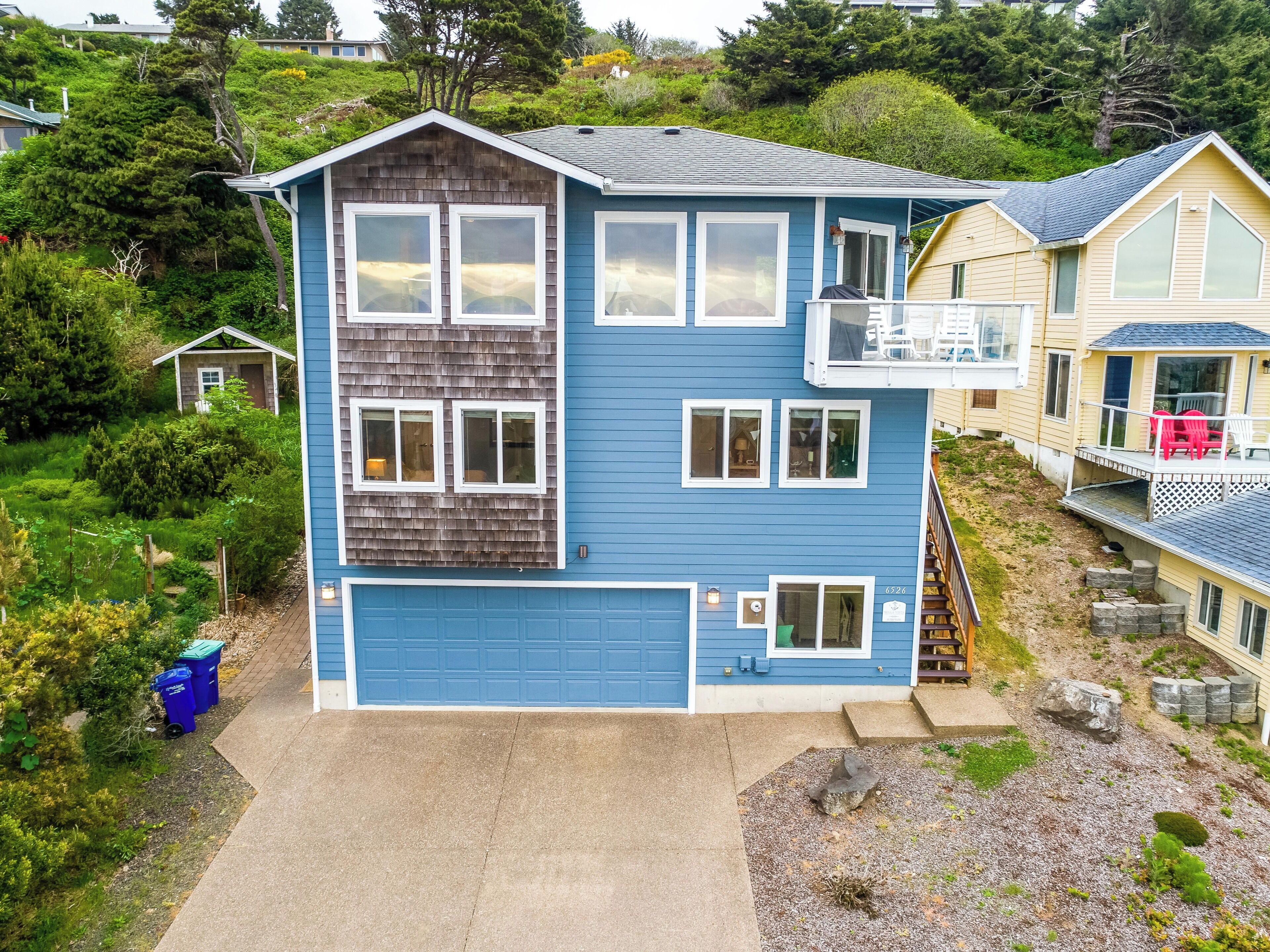 Une maison à deux étages avec une porte de garage bleue, un balcon et un petit abri de jardin à l’arrière.