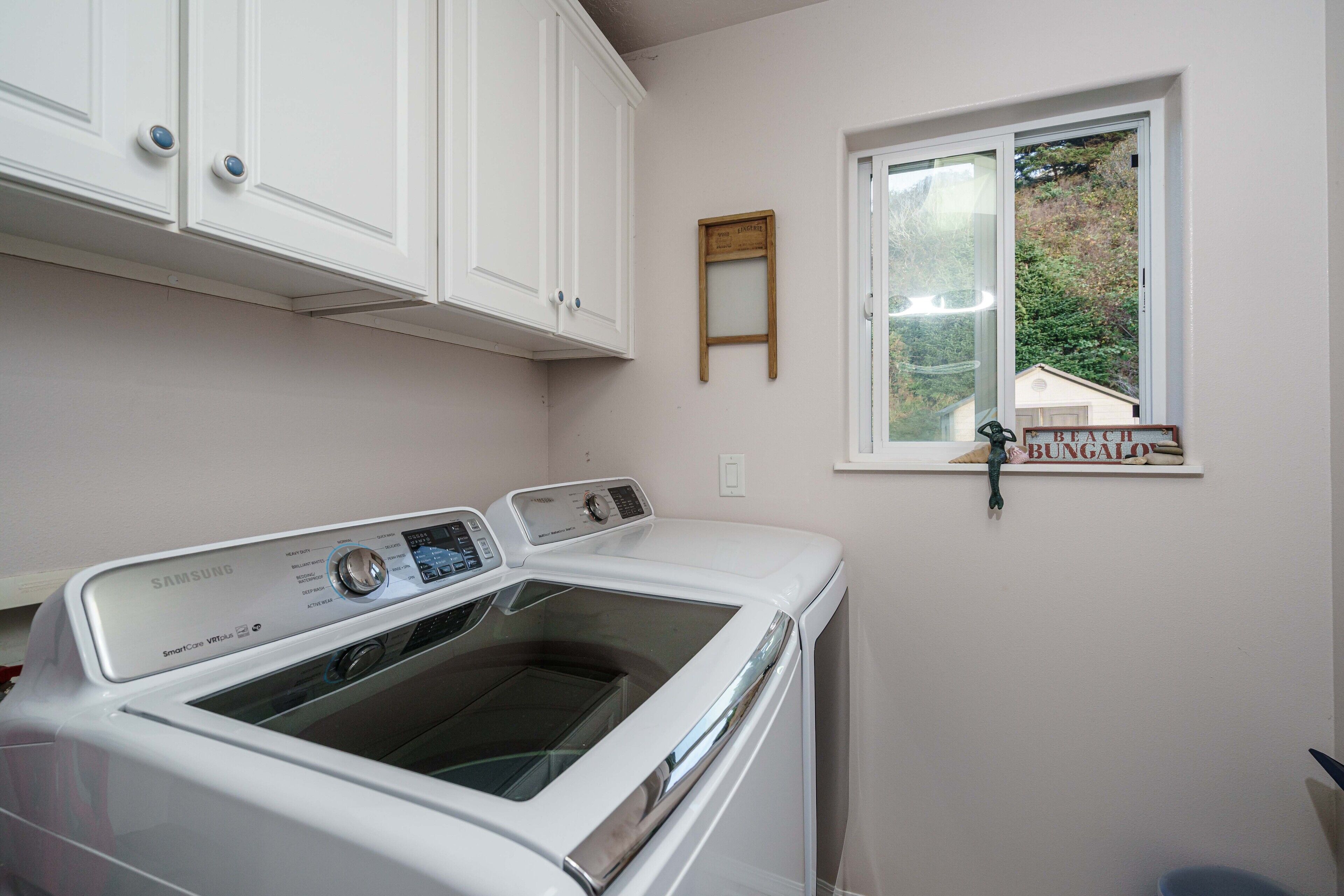 A laundry room with a front-loading washing machine, white cabinets, a window with a view of a house, and a small wooden sign on the window sill.