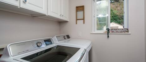 A laundry room with a front-loading washing machine, white cabinets, a window with a view of a house, and a small wooden sign on the window sill.