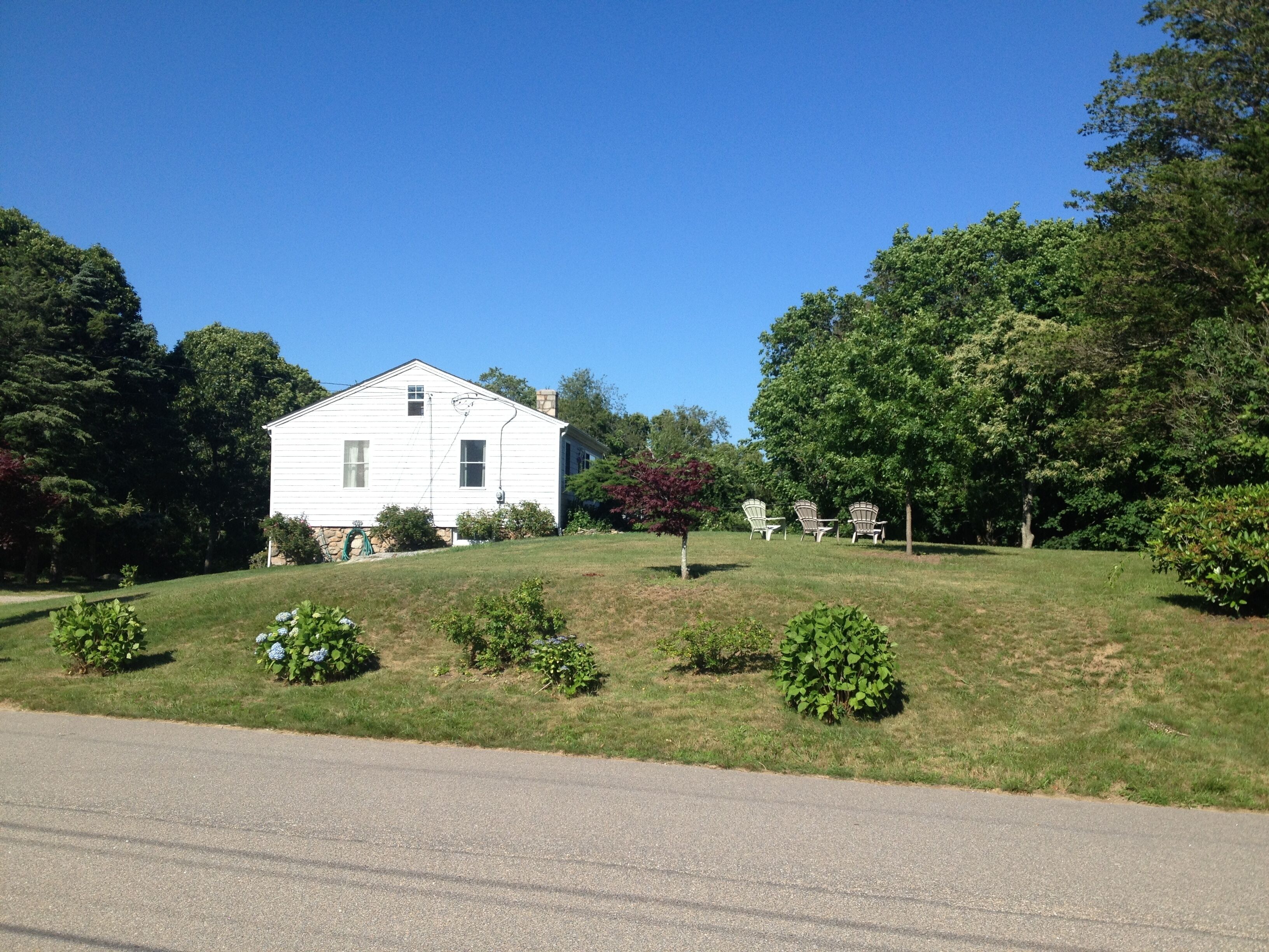 Une maison blanche à deux étages, avec une pelouse devant, un petit jardin et deux chaises d’extérieur.