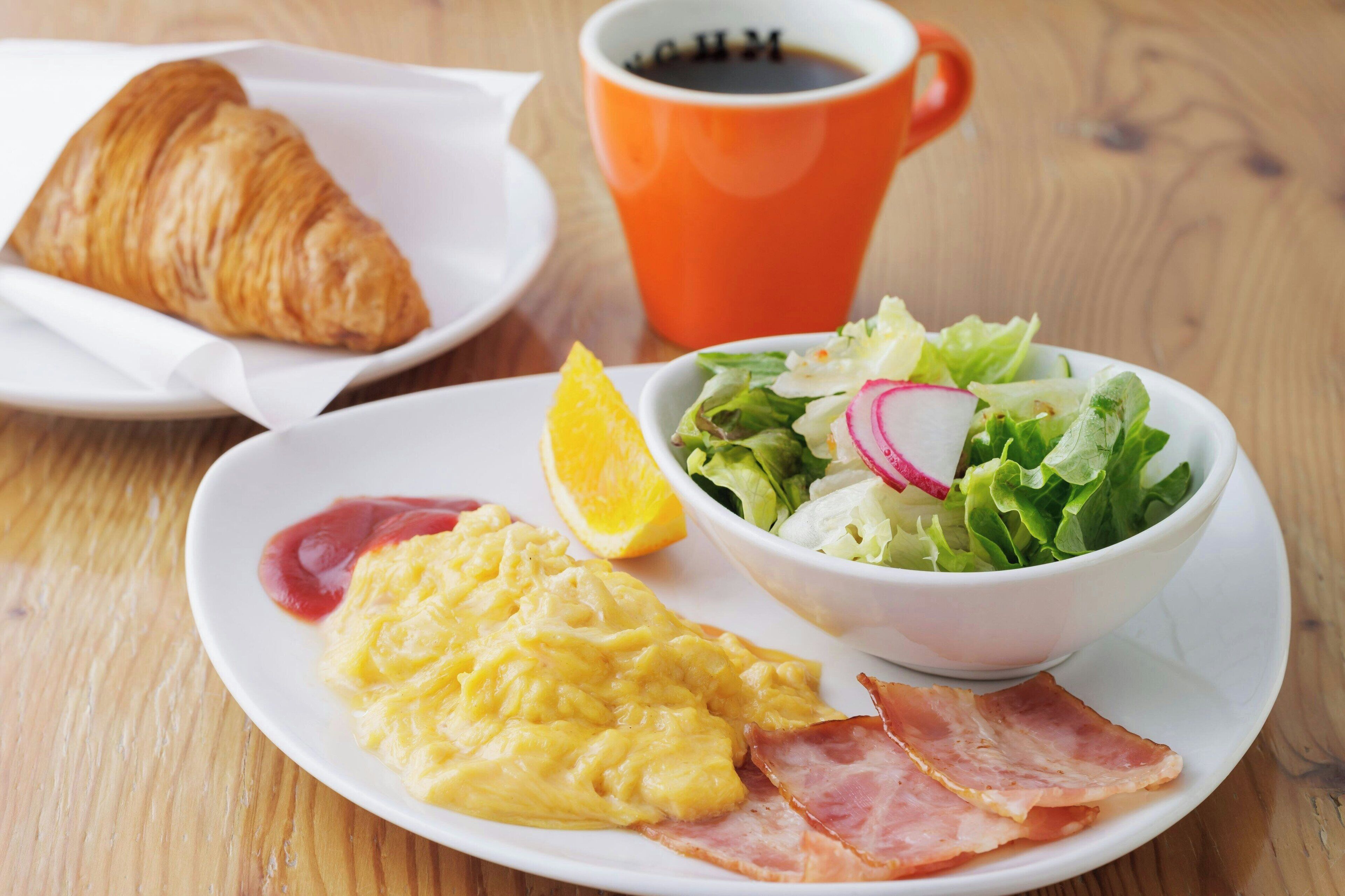 Un plat amb ous remenats, bacon, un croissant, un bol de amanida amb rave i una làmina de taronja.