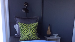 Un lit recouvert d’une couvre-lit blanche et bleue, un oreiller à motifs verts et noirs, une table de chevet avec une lampe, et une fenêtre avec des rideaux.
