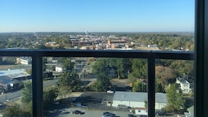 Une vue panoramique sur une ville, prise depuis un point d’observation élevé, qui présente un mélange de bâtiments commerciaux et résidentiels, un parking et un ciel bleu dégagé.