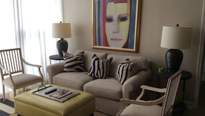 Un salon avec un canapé beige, deux fauteuils, un pouf jaune et un tableau abstrait encadré accroché au mur.