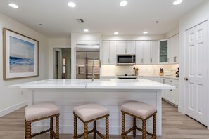 A modern kitchen with white cabinetry, a large island with bar stools, stainless steel appliances, and recessed ceiling lights.