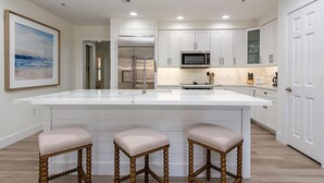 Cozinha moderna com armários brancos, uma ilha grande com banquetas, eletrodomésticos de aço inoxidável e iluminação embutida no teto.
