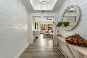Un couloir aux murs recouverts de panneaux blancs, doté d’un plancher en bois, d’un ventilateur de plafond, d’un miroir rond et d’une plante.