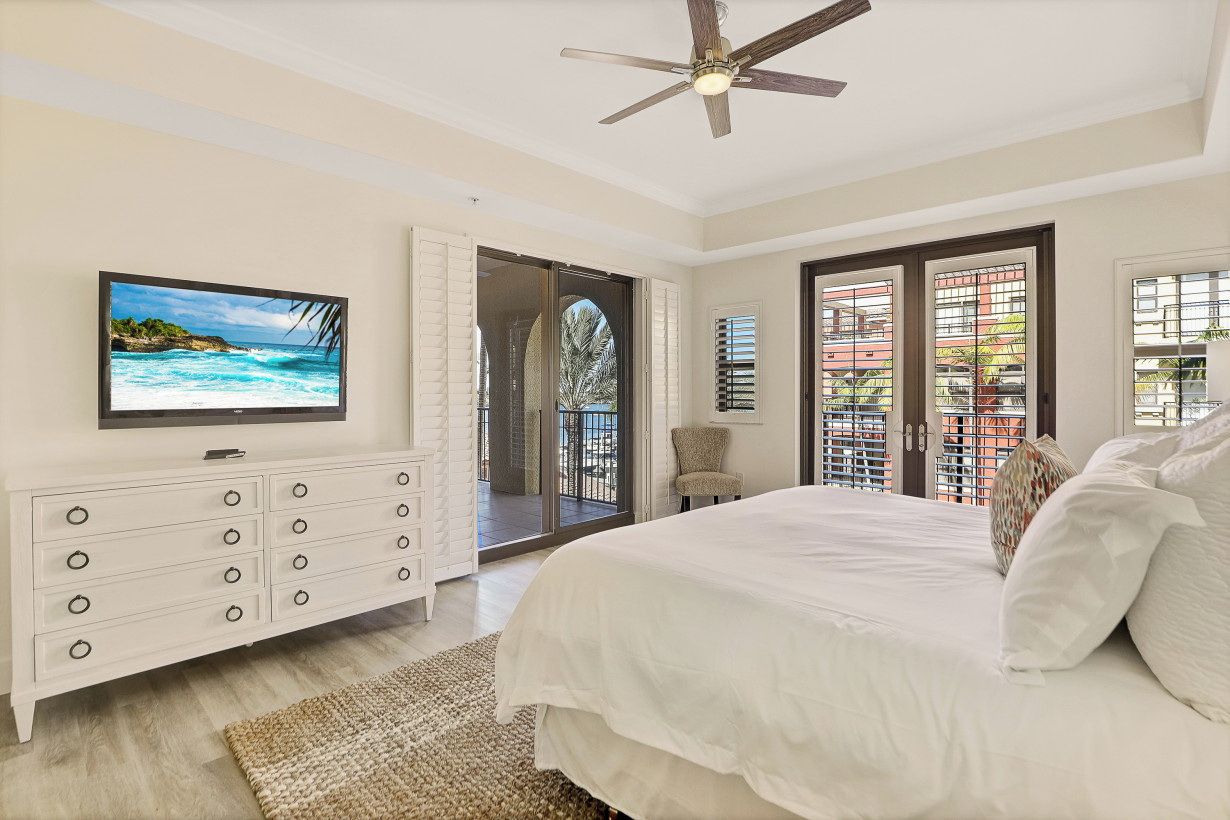A bedroom with a large bed, a television on the wall, a ceiling fan, and a view of the outside balcony.