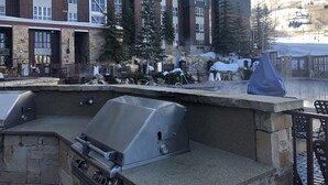 En udendørs terrasse med grill, stenvægge og udsigt til en høj bygning med stolelift i baggrunden.