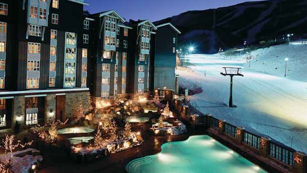 Et hotel med flere etager, et skiområde i baggrunden og et oplyst poolområde.