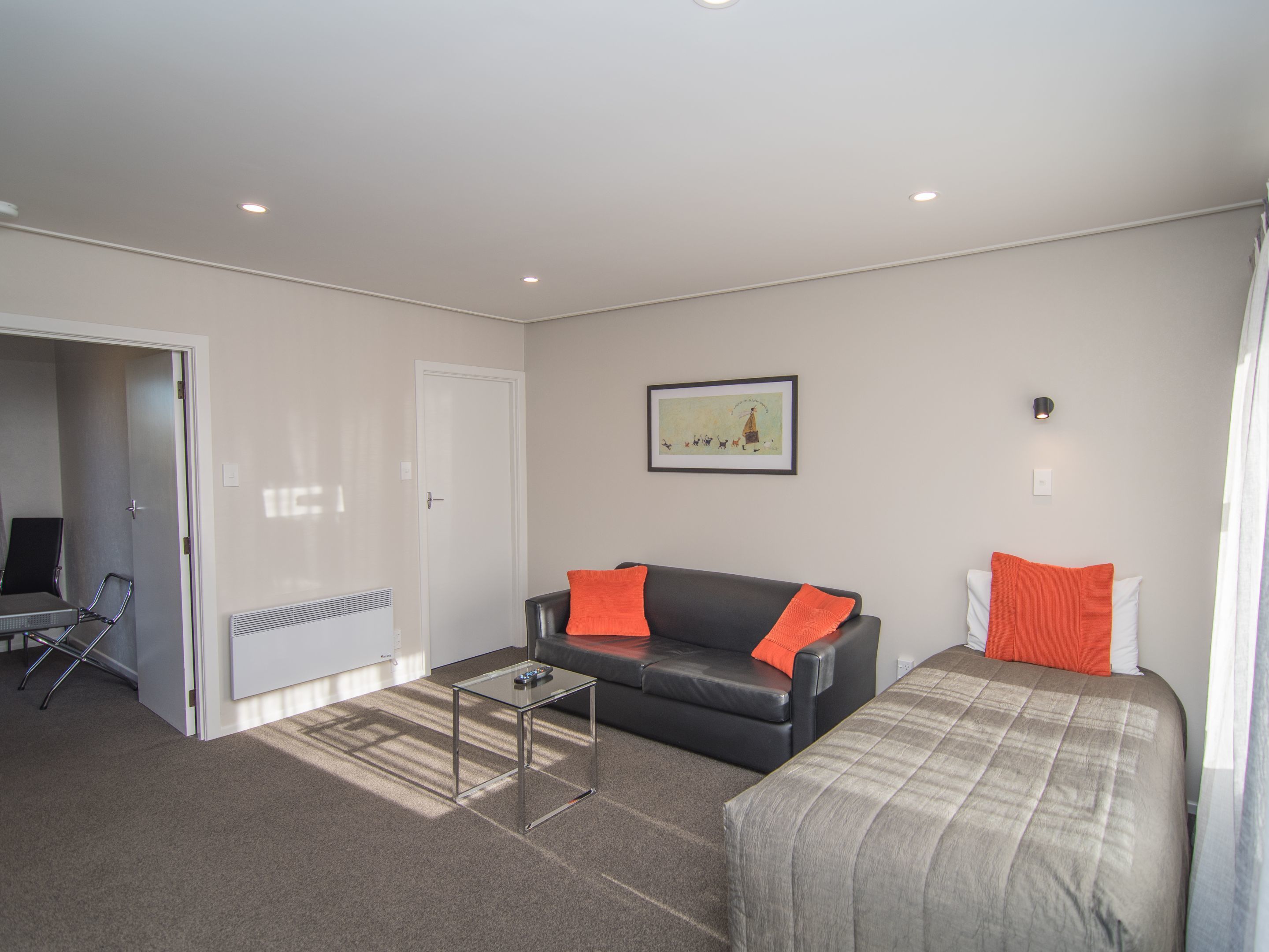 A modern, well-lit room with a black leather sofa, a glass coffee table, and a bed with a grey and orange bedspread.