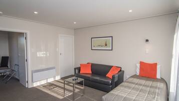 A modern, well-lit room with a black leather sofa, a glass coffee table, and a bed with a grey and orange bedspread.