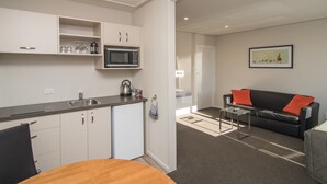 A compact kitchen with a microwave, sink, and cabinets. There is a dining table with chairs and a black leather sofa with orange cushions. A framed picture hangs on the wall. - Cedar Lodge Motel (Timaru)