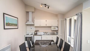 A dining area with a black table, white chairs, a framed picture on the wall, and a kitchen in the background. - Cedar Lodge Motel (Timaru)