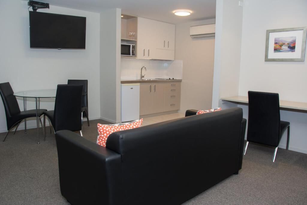 A compact living space with a sofa, a dining table, chairs, a kitchenette, and a flat-screen TV mounted on the wall.