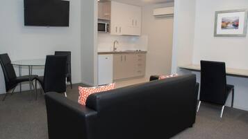 A compact living space with a sofa, a dining table, chairs, a kitchenette, and a flat-screen TV mounted on the wall.
