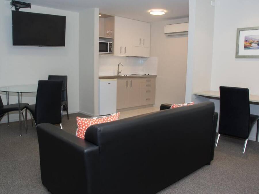 A compact living space with a sofa, a dining table, chairs, a kitchenette, and a flat-screen TV mounted on the wall.